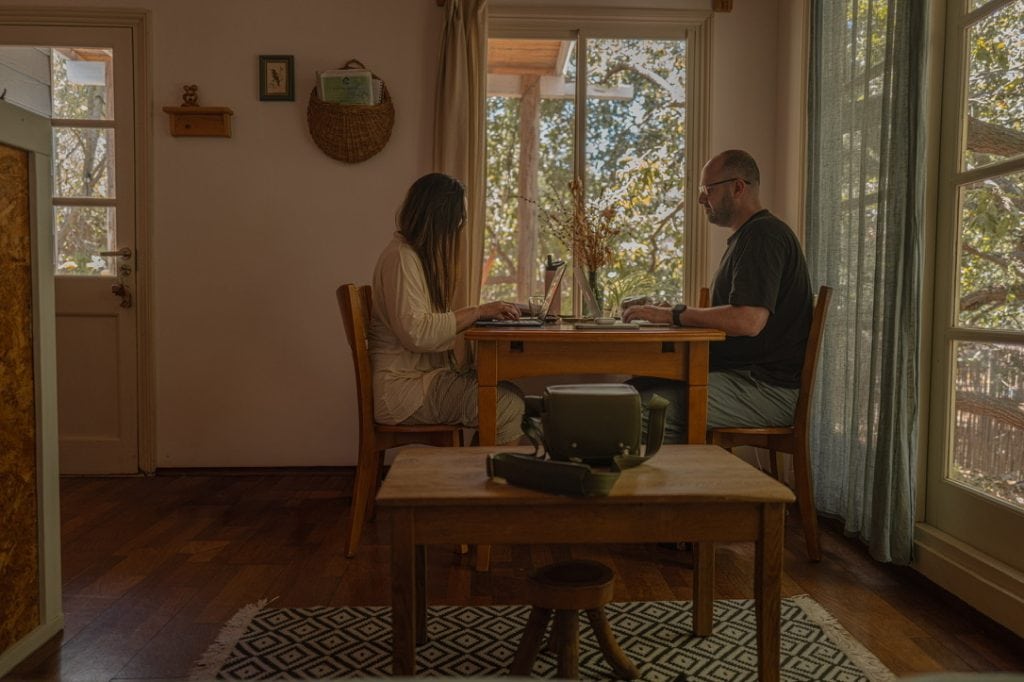 עומר ברק כותב מול מחשב ליד שולחן עץ, מול רותם, בחדר מואר בחלונות